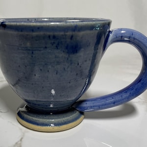 May include: A blue ceramic mug with a wide base and a rounded handle. The mug has a speckled glaze and a light brown rim.
