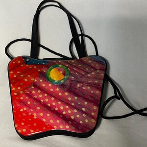 May include: A small, colorful handbag with a unique design. The bag features a red and pink patterned surface with gold polka dots and a black trim. It has black handles and a long black strap for shoulder or crossbody wear.