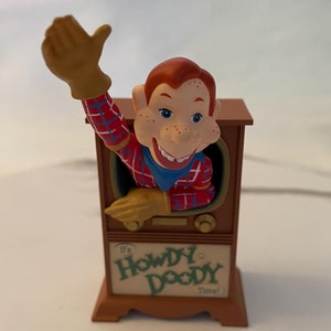May include: A brown wooden television set with a red-haired puppet with a red and blue plaid shirt sticking out of the screen. The television has a green label that reads "It's Howdy Doody Time!"