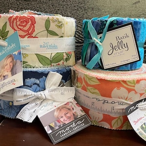 May include: Stacks of fabric rolls in various floral patterns, including roses and blue flowers, are tied with ribbons and tags. One roll is labeled "Batik Jelly" with dimensions of 2.5" x 22". The fabrics are in shades of blue, orange, and green.