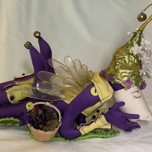 May include: A whimsical fairy doll with purple and yellow accents, resting on a green leaf-shaped base. The fairy has a crescent moon-shaped face, clear wings, and a gold-colored hat adorned with flowers. A small basket of berries sits nearby.