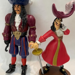 May include: Two Captain Hook figurines. One wears a burgundy jacket, blue pants, and a blue hat. The other is in a red coat and a burgundy hat with a feather, holding a sword. Both have black boots.