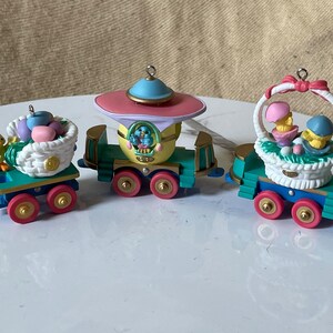 May include: A three-piece Easter train ornament set with a bluebird, chicks, and baskets of eggs. The train is green, yellow, blue, and pink.
