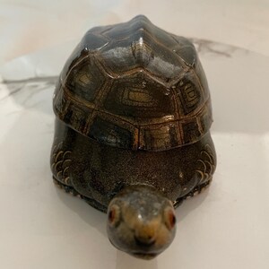 May include: A small, ceramic turtle figurine with a dark brown and gold shell. The turtle has a detailed face with red eyes and a small, upturned nose.