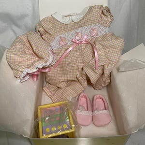 May include: A baby doll outfit set in a box. The set includes a pink and yellow plaid romper with white lace trim, pink satin ribbon, and matching pink slippers. A small toy is also included.