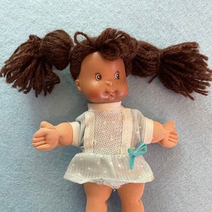 May include: A small brown-skinned doll with brown yarn hair in pigtails wearing a light blue dress with white lace and a turquoise bow.