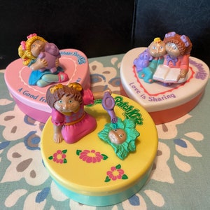 May include: Three heart-shaped boxes with different colored lids and characters from the cartoon show "The Care Bears" on top. The boxes are labeled "A Good Friend", "Love is Sharing", and "Patch Kids".