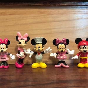 May include: Five small plastic figurines of Mickey Mouse and Minnie Mouse characters. The characters are wearing different outfits, including a pink dress, a red and yellow jumpsuit, and a white and gray chef's outfit.
