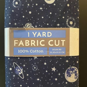 May include: A dark blue fabric cut with a celestial pattern of white stars, planets, and moons. The fabric is 36x44 inches and made of 100% cotton. The label reads "1 Yard Fabric Cut".