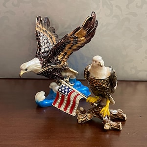 May include: A decorative figurine featuring two detailed eagles. One eagle is in flight, the other perched on a branch. Both are brown, gold, and white, with a small American flag. The base is blue.