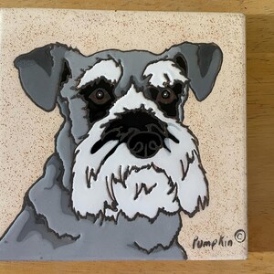 May include: A ceramic tile with a hand-painted image of a gray and white miniature schnauzer dog. The dog's face is the main focus of the image, and the tile is signed "Pumpkin" in the bottom right corner.