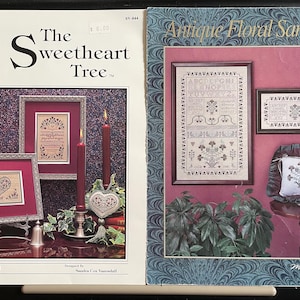 May include: Two cross-stitch patterns, one titled "The Sweetheart Tree" and the other titled "Antique Floral Sampler". Both patterns feature floral designs and text. The Sweetheart Tree pattern includes a heart and the text "Treasured Trio - II". The Antique Floral Sampler pattern includes an alphabet and the text "ABCDE FGH IJKLM NOPQ RSTUVWXYZ".