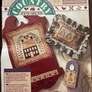 May include: A country-themed craft kit with cut-outs for creating home decor. The kit includes a wooden sign with the text "Warm Hearth Open House Est. 1906", a pillow with the text "Three Oaks Farm", and a small clock with a pineapple design. The kit also includes additional panels and craft cut fabrics.