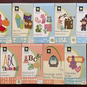 May include: A collection of Cricut cartridges in various designs. The cartridges feature themes like Doodlecharms, Wild Card, Cindy Loo, Winter Woodland, Paper Doll Dress Up, Jubilee, Storybook, George and Basic Shapes, and Fabulous Finds. Each cartridge has a unique design.