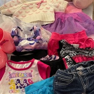 May include: A collection of clothing items for a doll or stuffed animal, including a pink sweater with a bow, a purple tulle skirt, a blue denim skirt, a pink and white shirt with the words "We Care" and Care Bears characters, a pink and black sequined skirt, a blue tulle skirt, a pink and white sweater, a pink and white tank top, a pink and white tutu, and a pair of pink boots.