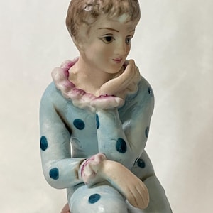 May include: A porcelain figurine of a seated figure in a light blue outfit with dark blue polka dots. The figure has short brown hair and is resting their chin on their hand. The collar is trimmed with pink ruffles.