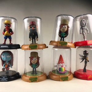 Puede incluir: Ocho figuras de personajes diferentes se muestran en c&uacute;pulas de pl&aacute;stico transparente. Los personajes incluyen Deadpool, Stan Pines, Mabel Pines, Bill Cipher, Dipper Pines, un gnomo, Sally y una abeja. Cada c&uacute;pula tiene una base con el nombre del programa del personaje impreso en ella.
