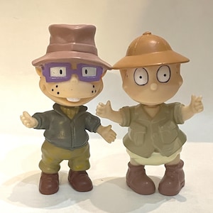 Op de afbeelding: Twee plastic speelfiguren. De linkerfiguur draagt een mauve hoed, een paarse bril, een donkergrijs jasje en een olijfgroene broek. De rechterfiguur draagt een beige tropenhelm, een lichtbeige overhemd en bruine laarzen met gespreide armen.