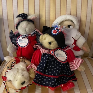 May include: Three plush toys dressed in red, white, and blue patriotic outfits. A white bunny wearing a top hat, a bear wearing a dress with stars, and a white dog wearing a hat. The toys are sitting on a striped chair.