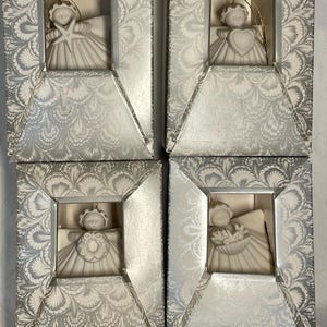 May include: Four white porcelain angel ornaments in individual boxes with silver and white patterned paper. Each angel is holding a different symbol: a starfish, a heart, a wreath, and a basket of flowers. The boxes have the text "Margaret  Fording" printed on them.