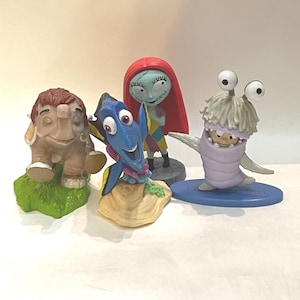Può includere: Una collezione di quattro piccole statuette di personaggi che include una creatura marrone simile a un elefante su erba verde, un pesce blu su una base color sabbia, una figura dalla pelle pallida con lunghi capelli rossi e una creatura viola chiaro.