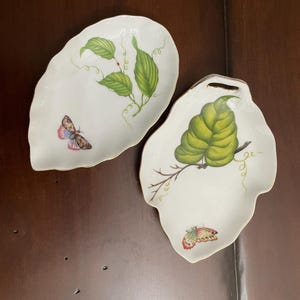 May include: Two white porcelain dishes shaped like leaves with scalloped edges and gold trim. One dish features a green leaf branch and a small butterfly. The other dish displays a larger green leaf and a colorful butterfly with pink and blue wings.