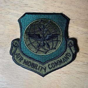 May include: A shield-shaped embroidered patch in shades of dark green and black. The center features a globe with a winged emblem. A banner at the bottom contains the text AIR MOBILITY COMMAND in black capital letters against a green background.