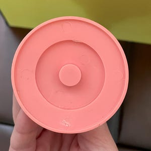 以下が含まれることがあります： 円形のデザインの、コーラル色のプラスチック製のオブジェクトのクローズアップ。オブジェクトは、隆起した中央部と、小さな円形のくぼみのある同心円状のリングを持っています。オブジェクトは手で持たれています。