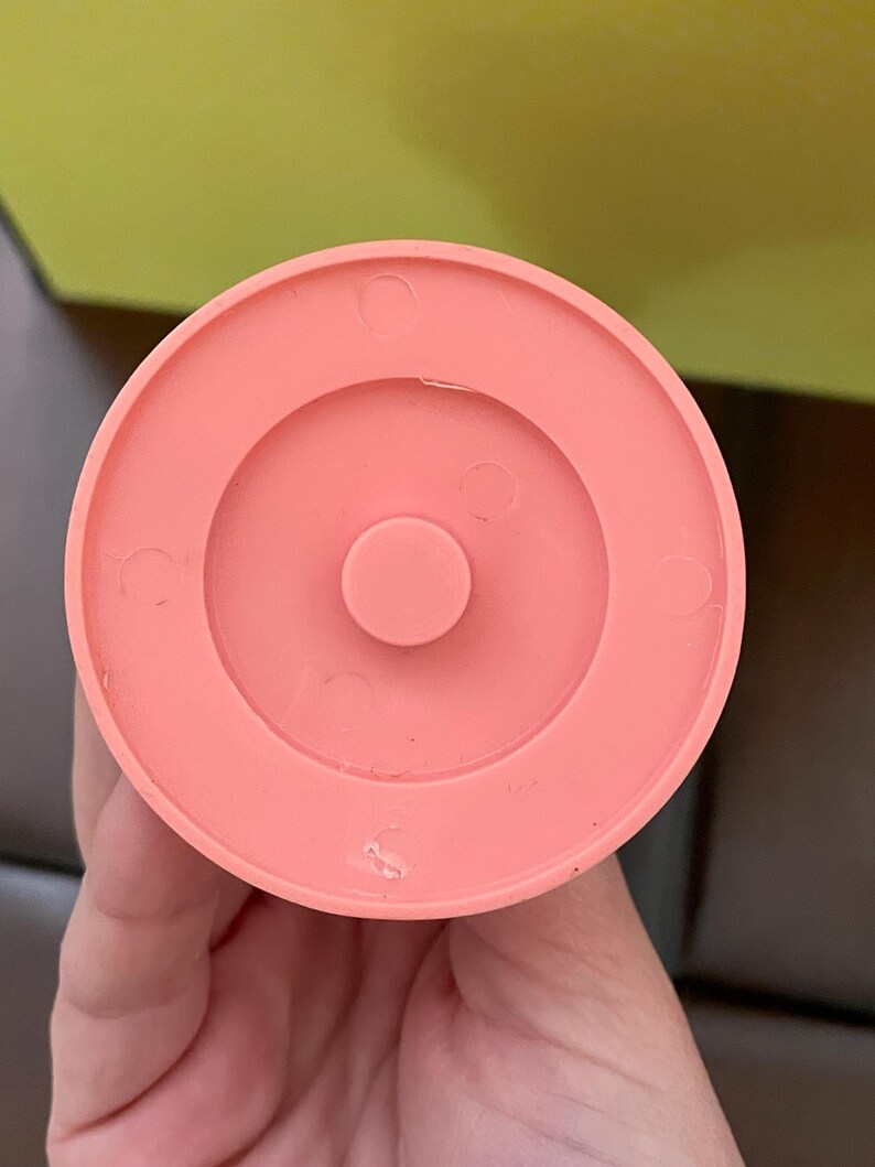 以下が含まれることがあります： 円形のデザインの、コーラル色のプラスチック製のオブジェクトのクローズアップ。オブジェクトは、隆起した中央部と、小さな円形のくぼみのある同心円状のリングを持っています。オブジェクトは手で持たれています。