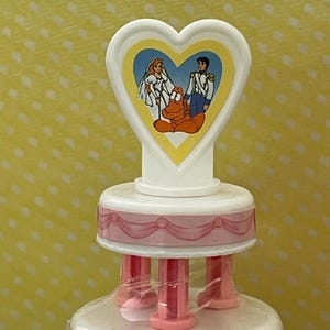 以下が含まれることがあります： 白とピンクのケーキトッパー。ハート型のデザインで、花嫁、新郎、赤いカニの漫画の画像が描かれています。ケーキトッパーは、ピンクのリボンとサポートを備えた多層設計になっています。