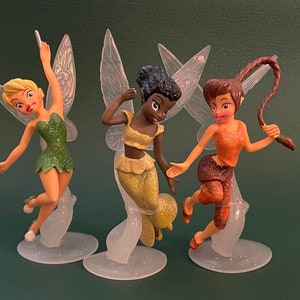 May include: Three fairy figurines with translucent wings and glitter accents. The fairies are standing on clear plastic bases. The fairies are wearing different colored outfits: green, yellow, and orange.
