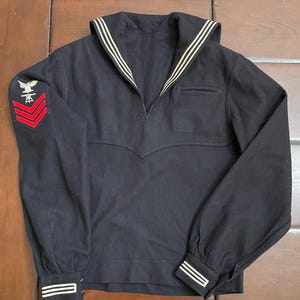 May include: A dark navy blue long-sleeve sailor jumper featuring a wide collar with three white stripes, a V-neckline, and cuffs with three white stripes. The right sleeve displays an embroidered eagle patch above three red chevron stripes.