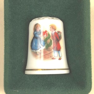 May include: A white porcelain thimble with a gold band at the base, depicting two children exchanging gifts. The thimble is presented in a dark green velvet box, offering a classic and elegant display.