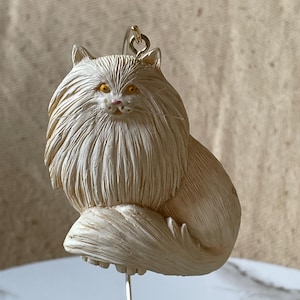 May include: White ceramic ornament of a fluffy cat with yellow eyes. The cat is sitting with its tail curled around its body. The ornament is attached to a gold stand.
