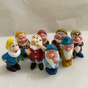 May include: A collection of eight small dwarf figurines, each wearing a distinct colored hat and tunic. The figures feature painted facial expressions, white beards, and varied poses, standing together in a group with colorful clothing details.