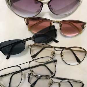 May include: A collection of six pairs of eyeglasses and sunglasses, including a pair of white sunglasses with the word "GUCCI" on the frame, a pair of brown sunglasses, a pair of black sunglasses, a pair of clear-framed glasses, a pair of silver-framed glasses, and a pair of silver-framed glasses with the text "FRATELLI LOZZA DESIGN" on the frame.