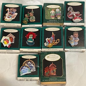 May include: A collection of miniature Keepsake ornaments in their original boxes. The ornaments feature various designs, including angels, swans, and holiday-themed figures. The boxes are green with gold accents.