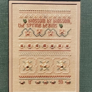 May include: A framed cross-stitch sampler with the text "blossom by blossom, the spring beams." The needlework features floral and geometric patterns in shades of pink, green, and brown on a beige background. The frame is a light beige color.