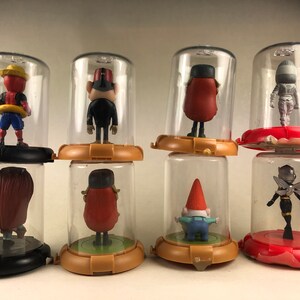 Puede incluir: Ocho peque&ntilde;as figuritas de pl&aacute;stico se muestran en c&uacute;pulas de pl&aacute;stico transparente sobre un fondo blanco. Las figuritas son de varios personajes, incluyendo un gnomo, una abeja, un personaje de cabeza roja y un personaje con fez.