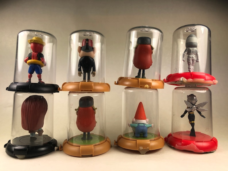 Puede incluir: Ocho peque&ntilde;as figuritas de pl&aacute;stico se muestran en c&uacute;pulas de pl&aacute;stico transparente sobre un fondo blanco. Las figuritas son de varios personajes, incluyendo un gnomo, una abeja, un personaje de cabeza roja y un personaje con fez.