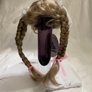 May include: A blonde doll wig with two braids tied with pink ribbons. The wig is styled with a bun on top and the braids are tied at the ends with pink elastic bands. The wig is displayed on a clear glass vase.