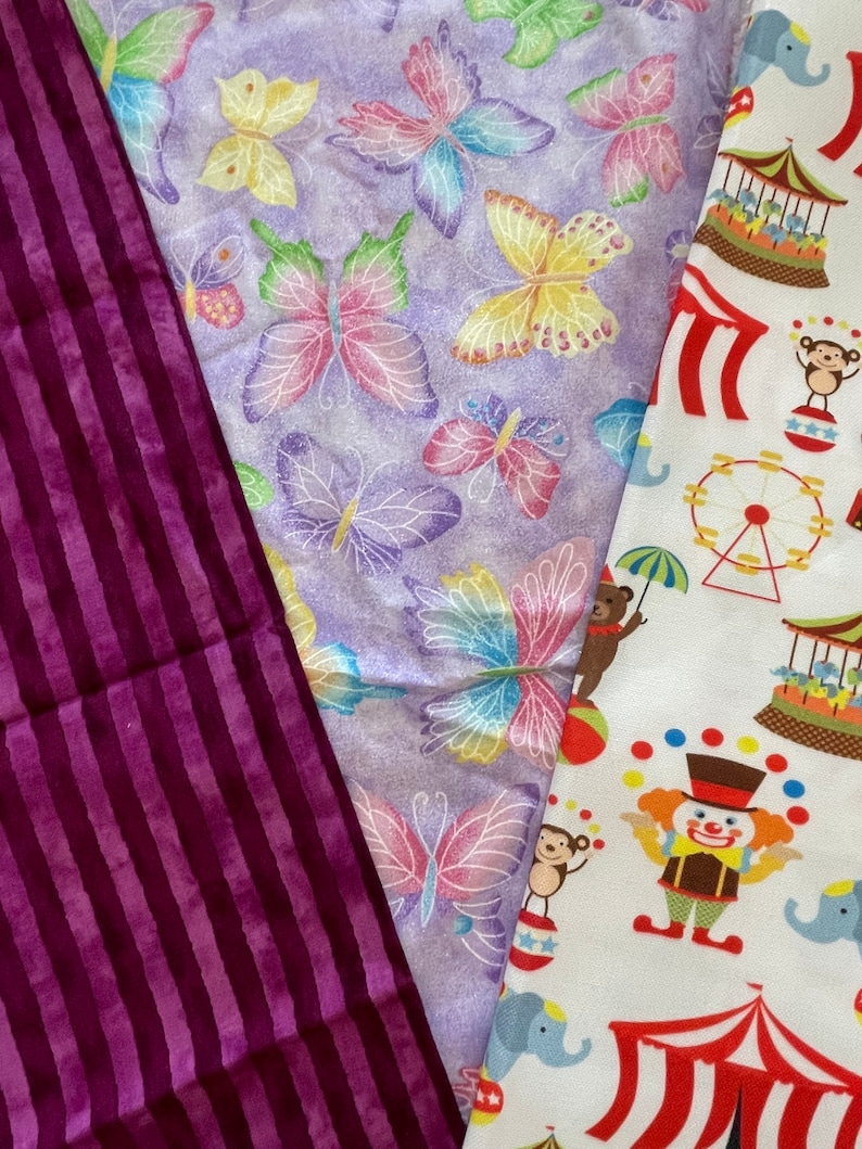 May include: Three fabric swatches, one with a purple and white striped pattern, one with a purple background and a repeating pattern of colourful butterflies, and one with a white background and a repeating pattern of circus-themed illustrations including a clown, an elephant, a Ferris wheel, and a tent.
