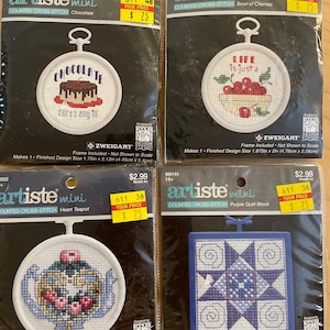 May include: Four "Artiste Mini" counted cross-stitch kits. Designs include a chocolate cake, a bowl of cherries, a heart teapot, and a purple quilt block. Each kit includes a frame. Finished design sizes vary from 1.75in x 2in to 2.5in x 2.125in.