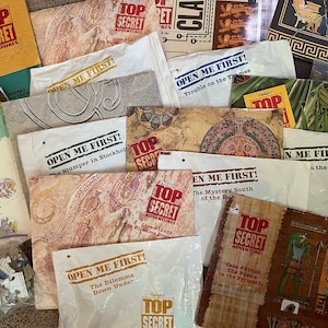 May include: A collection of Top Secret Adventures game envelopes with different titles and case numbers. The envelopes are made of paper and have a vintage look. The text on the envelopes includes "Open Me First!" and "Case #31682 The Puzzle in the Pyramids".
