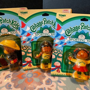 May include: Three Cabbage Patch Kids Sporting 'Kids figures on their packaging. The figures are a baseball player wearing a green and yellow uniform, a girl with a red skirt and yellow shirt holding a golf club, and a boy wearing a cowboy hat and holding a baseball bat.