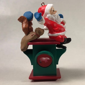 May include: A red and white Santa Claus ornament with a blue hat and a brown squirrel ornament on a green and red base. The base has a circular opening with a white pig silhouette.