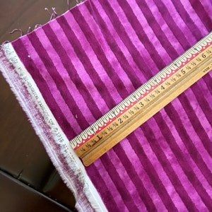 May include: Purple and white striped fabric with a wooden ruler measuring 15 cm. The ruler has markings for 1/4, 1/2, 3/4, and full inches.