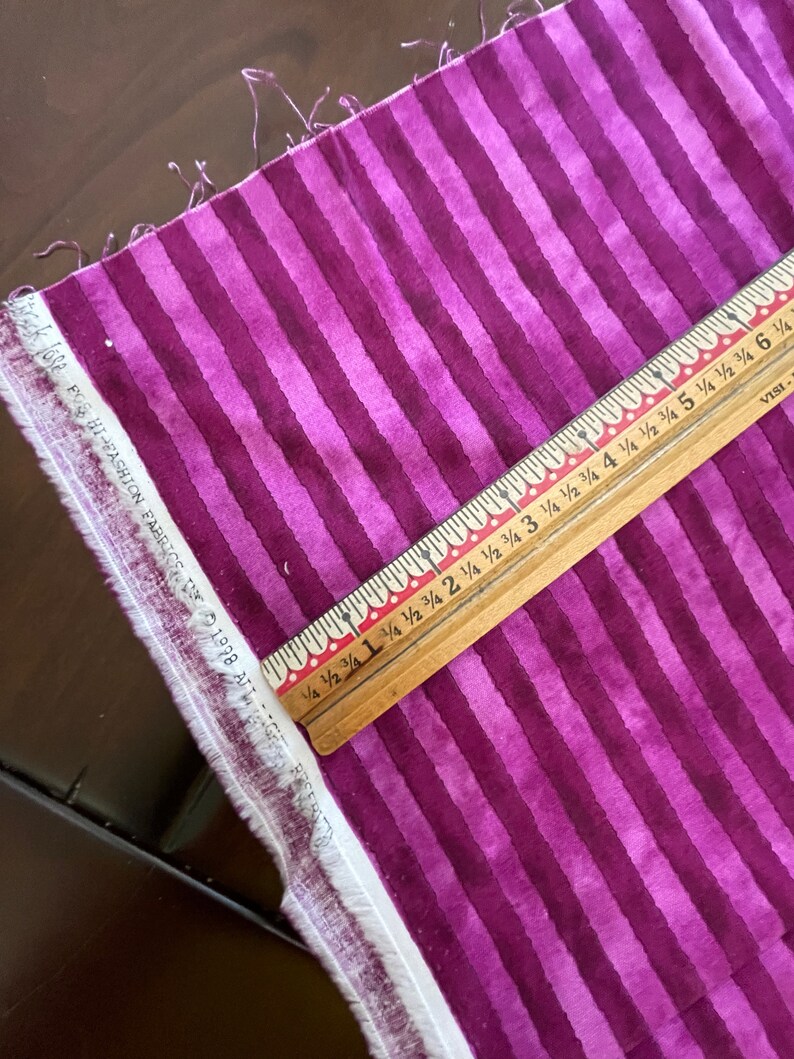 May include: Purple and white striped fabric with a wooden ruler measuring 15 cm. The ruler has markings for 1/4, 1/2, 3/4, and full inches.