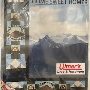 May include: A quilting pattern featuring a sea otter with the text "Home Sweet Homer" and a mountain landscape. The pattern is from Row by Row Experience 2016 and is by Cookin Graphics. The pattern is being sold at Ulmer's Drug & Hardware.
