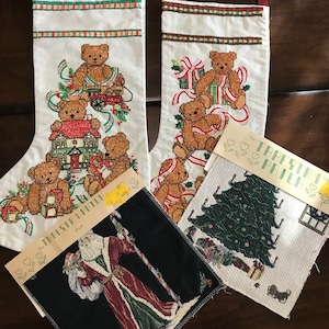 May include: Two white Christmas stockings with embroidered teddy bears and presents. A white and green Christmas tree tapestry with the text "The Tapestry Path" and a black tapestry with a Santa Claus figure.